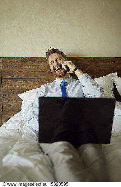 Businessman lying on bed in hotel room laughing on the phone