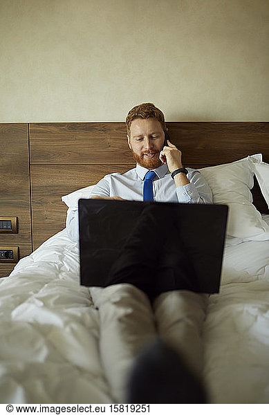 Businessman lying on bed in hotel room using laptop and cell phone
