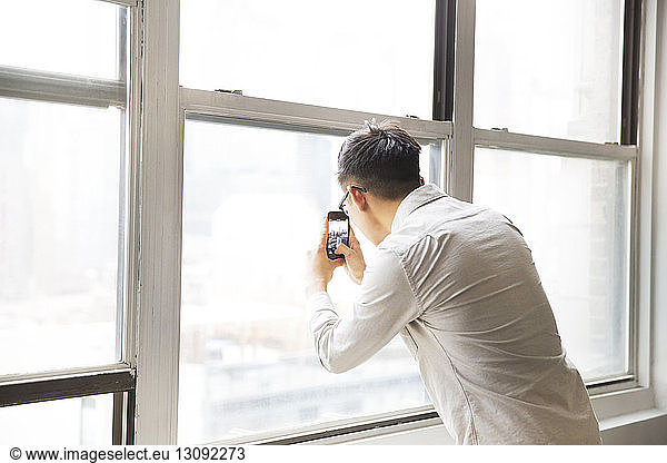 Businessman photographing from window through smart phone in creative office