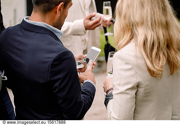 Businessman showing mobile phone to businesswoman while holding champagne flute