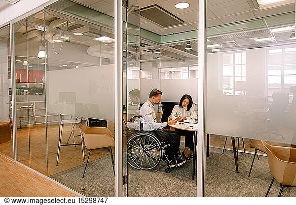Businessman sitting on wheelchair while working with female colleague in board room at work place seen through doorway