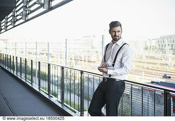 Businessman standing against railing and working on digital tablet  Munich  Bavaria  Germany