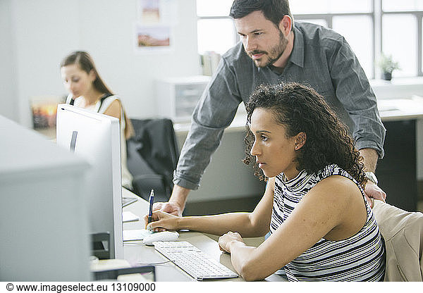 Businessman standing by female colleague using desktop computer at desk