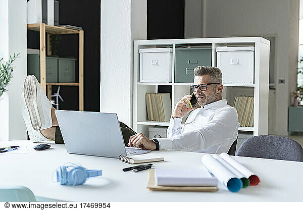 Businessman talking on smart phone while using laptop in office