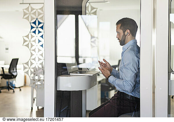 Businessman talking on video call over laptop while sitting in telephone booth at office