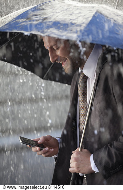 Businessman text messaging on cell phone under umbrella in rain