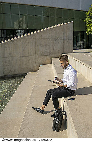 Businessman using digital tablet while sitting on steps by briefcase