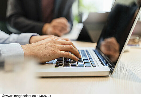 Businessman using laptop at desk with colleague in background