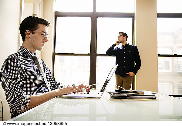 Businessman using laptop at table with colleague using phone in background