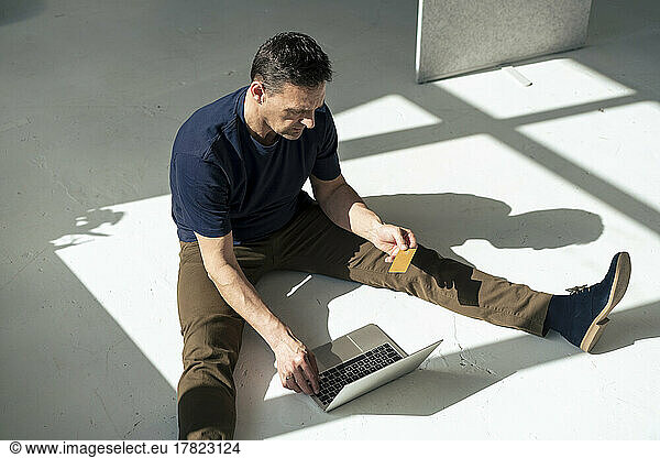 Businessman using laptop holding credit card sitting on floor at office