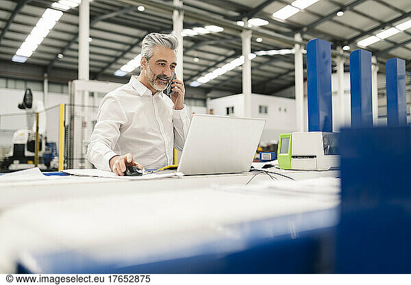 Businessman using laptop talking on smart phone in factory