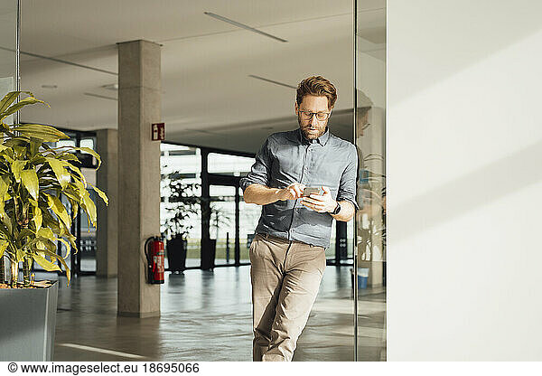 Businessman using mobile phone leaning on glass door at office lobby