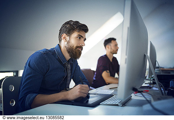 Businessman using pen tablet while working by colleague at desk in office