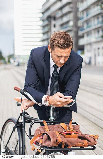 Businessman using smart phone in city while leaning on bicycle