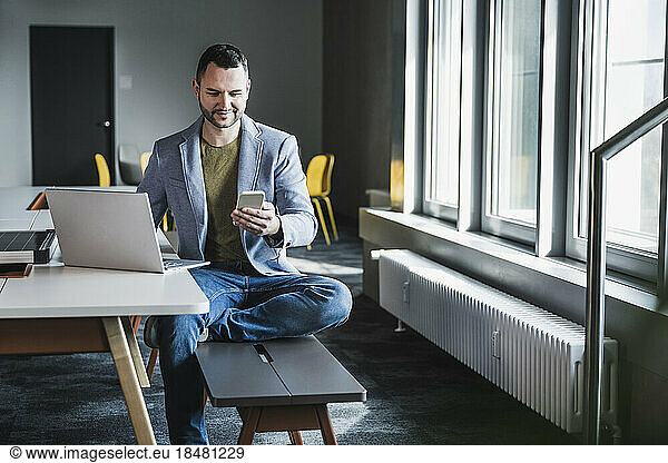 Businessman using smart phone sitting on bench by laptop at desk in office