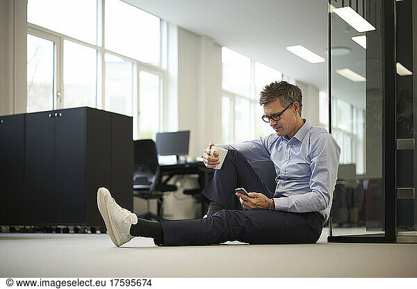 Businessman using smart phone sitting on ground in modern office