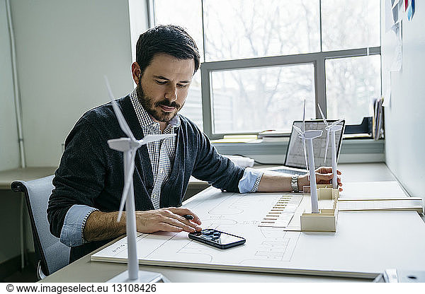 Businessman using smart phone while arranging wind turbine models on desk