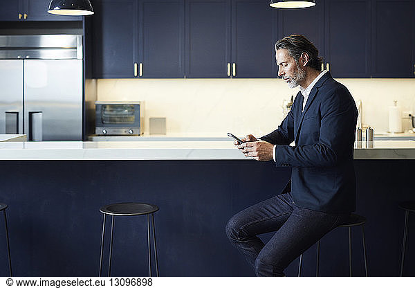 Businessman using smart phone while sitting on stool at kitchen counter in creative office