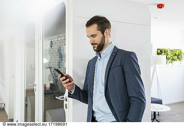 Businessman using smart phone while standing by telephone booth at office