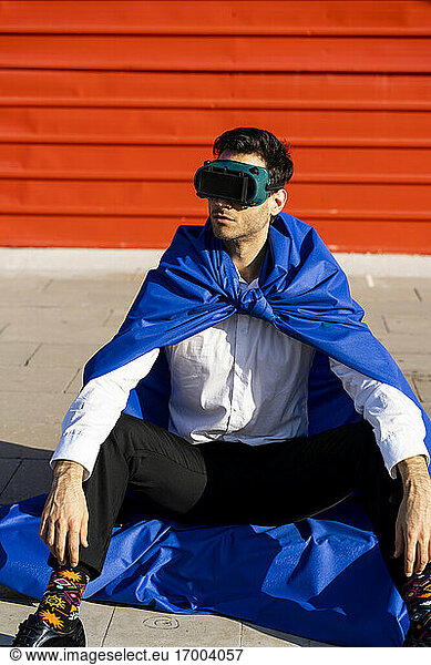 Businessman wearing superhero cape and VR goggles sitting on curb