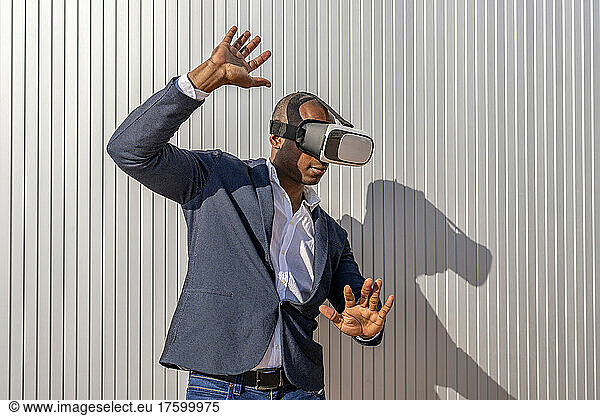 Businessman wearing virtual reality simulator gesturing in front of wall on sunny day