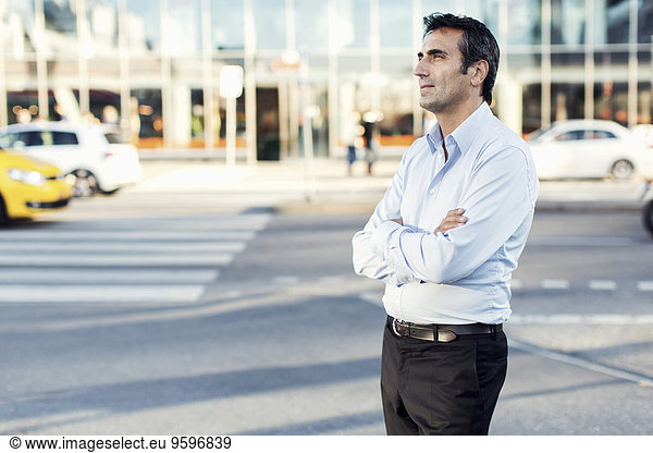 Businessman with arms crossed looking away while standing by city street