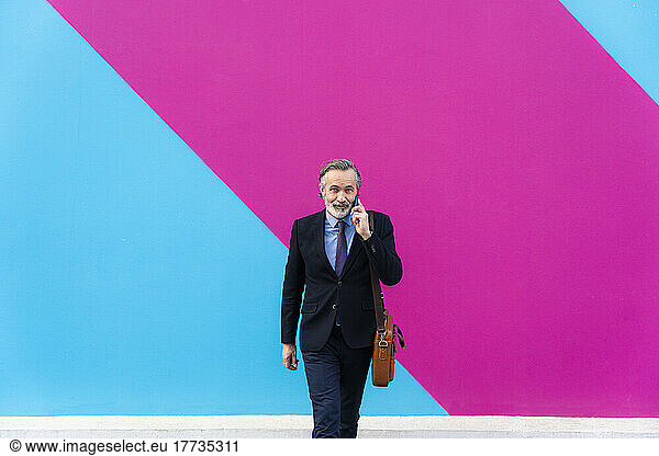 Businessman with bag talking on smart phone walking in front of pink and blue wall