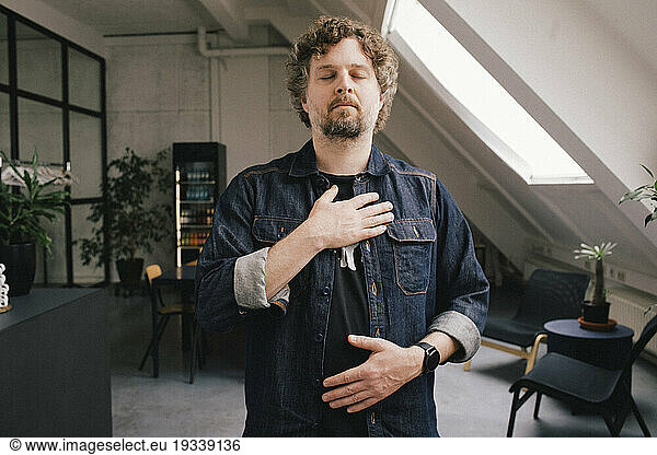 Businessman with eyes closed and hand on chest standing at office