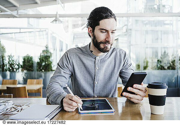 Businessman with graphics tablet and reusable coffee cup using smart phone in office