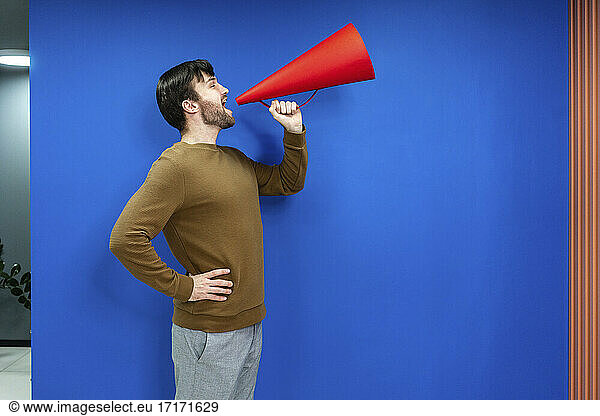 Businessman with hand on hip screaming in megaphone by blue wall at work place