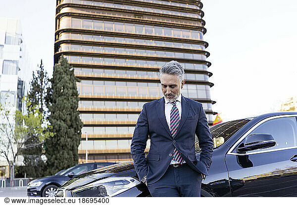 Businessman with hands in pockets looking down by car in front of building
