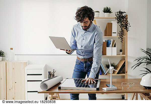 Businessman with laptop keeping hand on solar panel at desk in office