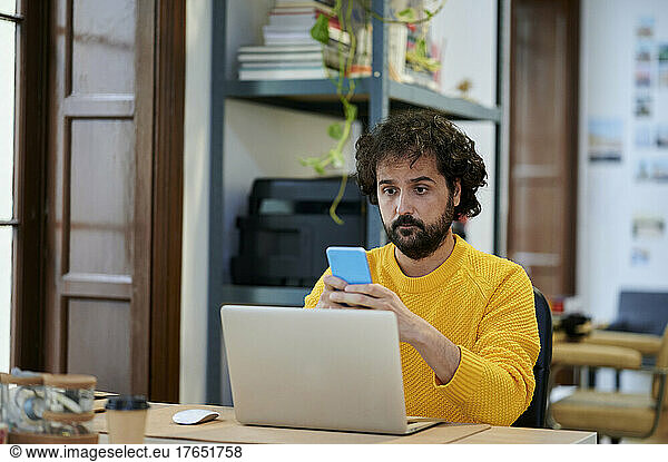 Businessman with laptop using smart phone at desk in office