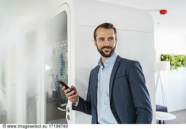 Businessman with mobile phone smiling while standing by telephone booth in office