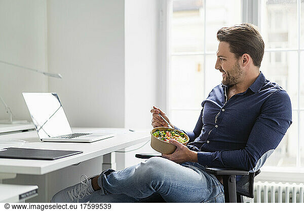 Businessman with salad bowl watching laptop at desk in office