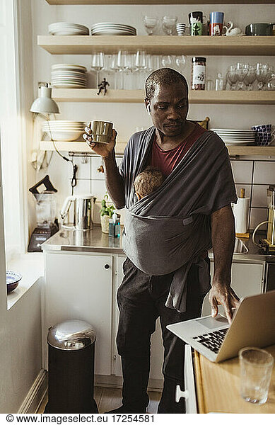 Businessman with son in baby carrier using laptop while holding cup at kitchen