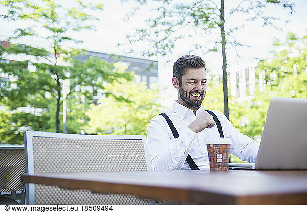Businessman working on laptop in outdoor cafe and smiling  Munich  Bavaria  Germany