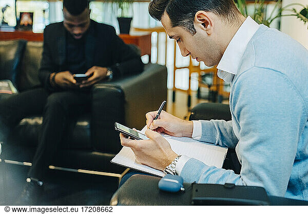 Businessman writing in book sitting by male colleague in office