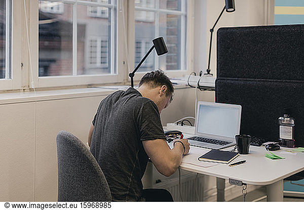Businessman writing while using laptop at desk in creative office