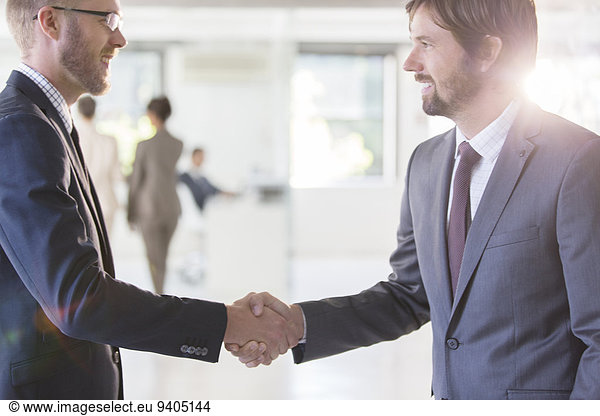 Businessmen wearing suits shaking hands in office  colleagues in background