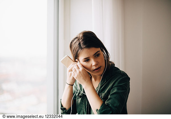 Businesswoman adjusting in-ear headphones while holding smart phone by window at creative office