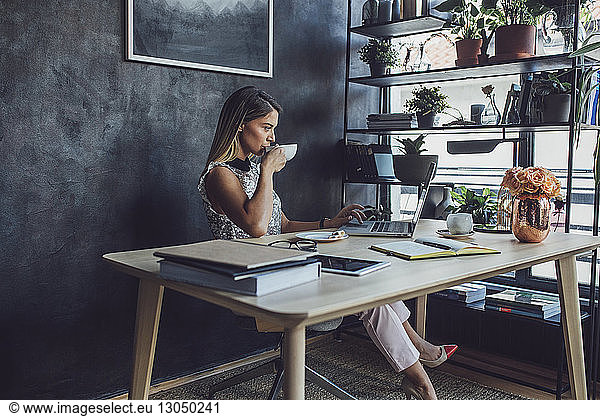Businesswoman drinking coffee while working at laptop computer in home office