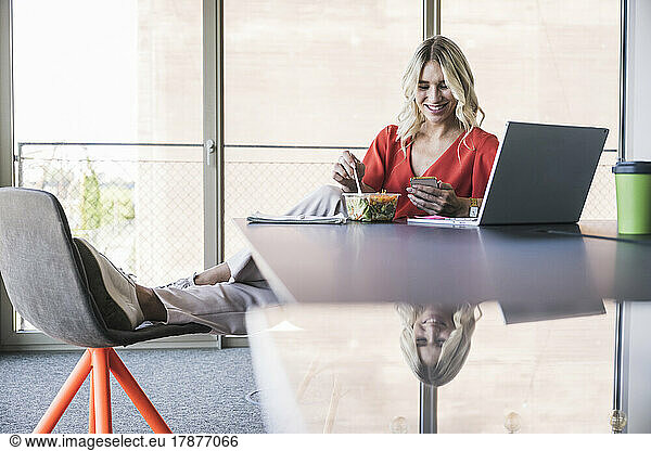 Businesswoman having lunch using smart phone sitting at desk in office