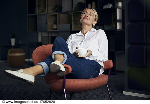 Businesswoman holding coffee cup sitting with eyes closed on chair in office