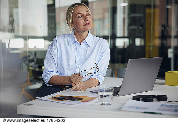 Businesswoman holding eyeglasses and laptop at desk in office