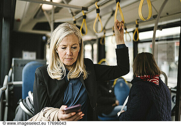 Businesswoman holding grab handle while using smart phone in bus