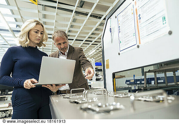 Businesswoman holding laptop with colleague pointing at machine in industry