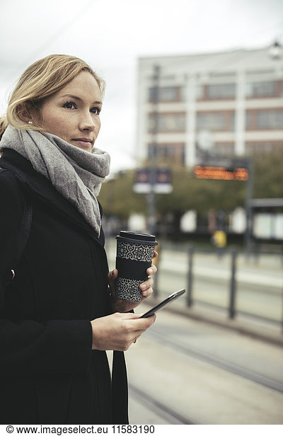 Businesswoman holding smart phone and disposable coffee cup at tram station