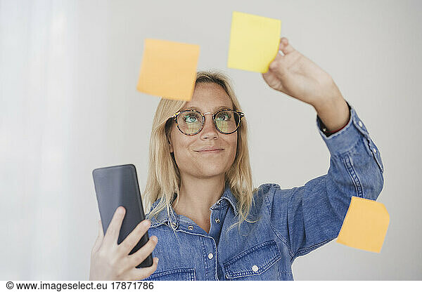 Businesswoman holding smart phone sticking adhesive notes on glass