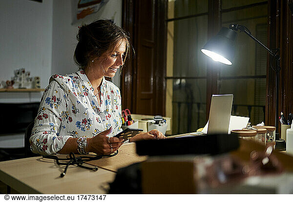 Businesswoman holding smart phone while working on laptop in office